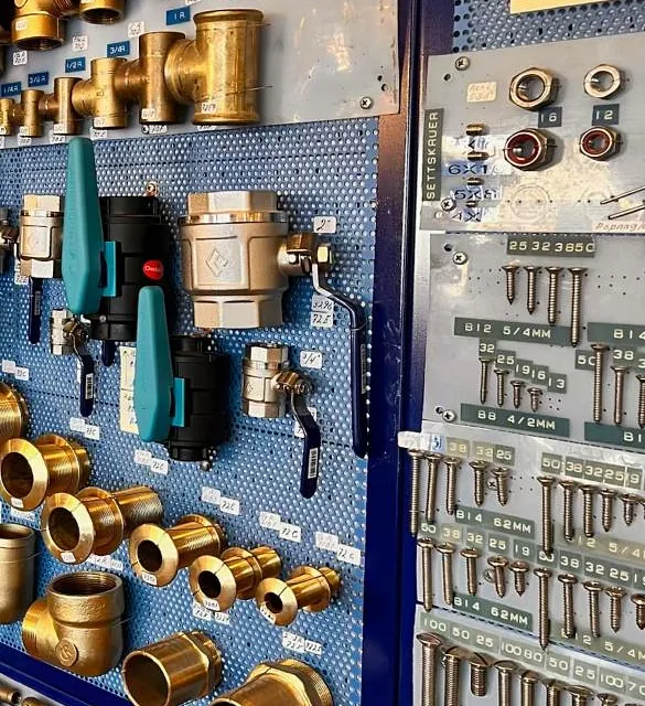 Vegg med ulike messing- og metallrørdeler, ventiler og skruer, merket med størrelser og priser.