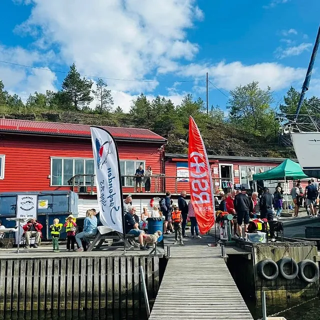 Folk samles ved en rød bygning i en småbåthavn, med flagg, boder og en stor seilbåt på land.